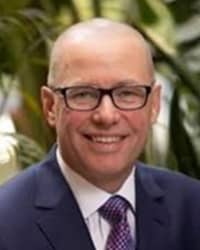 Matt Peterson wearing a dark blue jacket, white collared shirt, and blue patterned tie, stands outside in front of greenery and smiles at the camera.