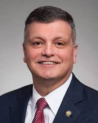 Noel J. Cárdenas, wearing a red tie, white collared shirt, and black suit coat, stands in front of a gray backdrop and smiles at the camera.
