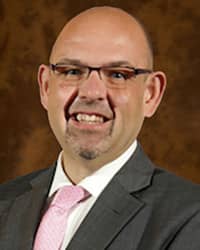 Rob Mach wearing a pink tie, white collared shirt, and gray suit coat, stands in front of a brown backdrop and smiles at the camera.