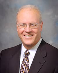 Mark Taylor, wearing glasses, a white collared shirt, a tie, and brown suit jacket, sits in his office and smiles at the camera.