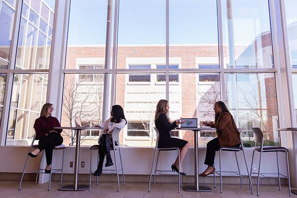 Four health administration students dressed professionally sit at tables in the Health Professions Building atrium. Two students sit with their legs crossed and converse with each other while the other two sit and talk at a different table with a laptop.
