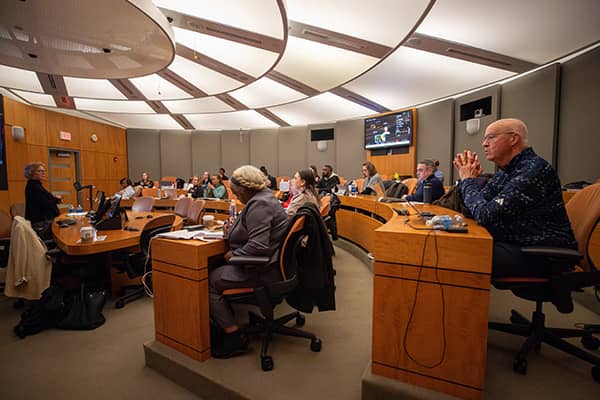 A group of Doctor of Health Administration students sit in two rows in the global telepresence room in the College of Health Professions Building listening to a speaking standing at the front of the room.