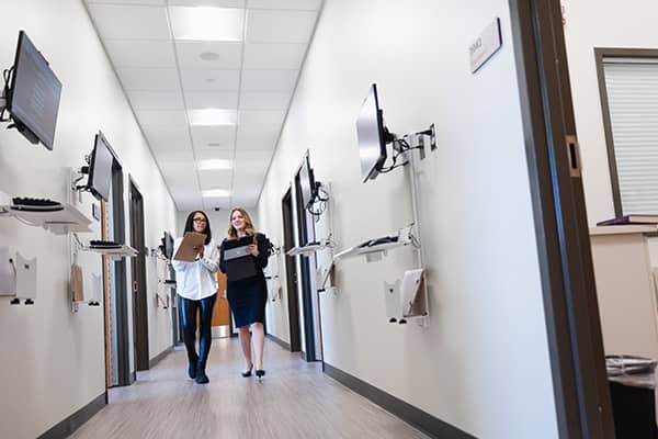 Two health administration students dressed professionally stand side-by-side holding clipboards and papers and walk down the hallway of the Interprofessional Education and Practice Center.