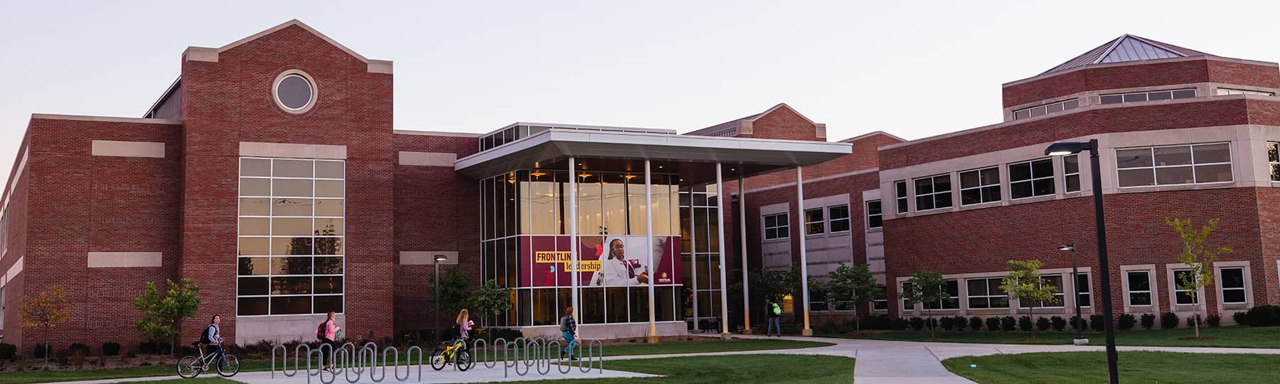 A photo of the south entrance of the two-story College of Health Professions building on the campus of Central Michigan University. There are sidewalks interconnected with a bike rack leading up to the class encased entrance supported by white columns.