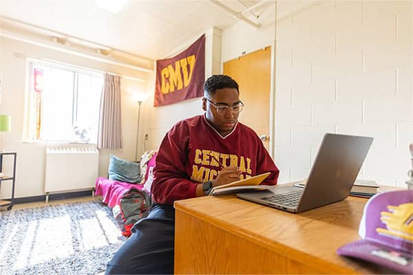 A student wearing glasses and a maroon Central Michigan pullover sits at a desk studying in his room in Emmons Hall. He is holding a pencil in his right hand writing in a notebook in front of a laptop positioned on his desk.
