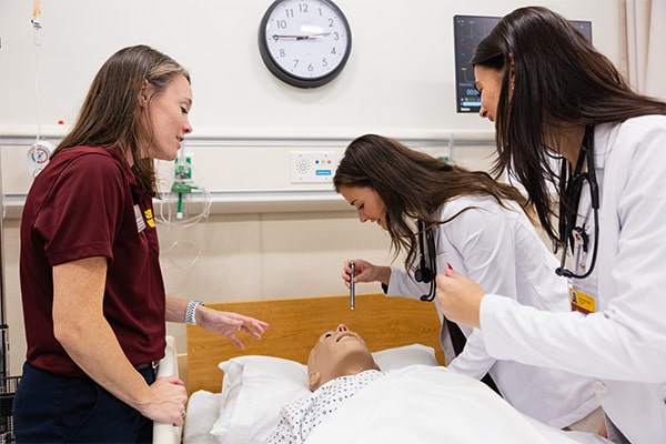 Faculty member Amy Malheim interacts with two students in white coats while working with a simulation mannequin in the IPEP Center.