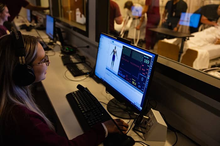 A student wearing headphones sits in the simulation control center at a desk with a computer.