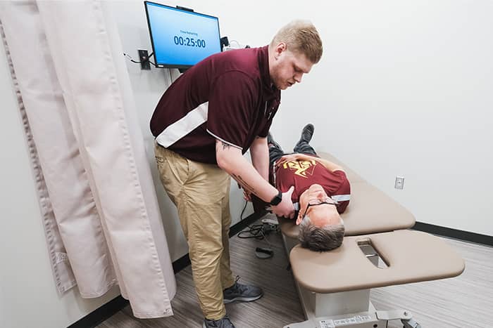 An athletic training student holds an older patient's shoulder and arm on an examination table.