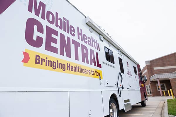 The white Mobile Health Central bus with Mobile Health Central painted in maroon and Bringing Healthcare to You in maroon on a yellow banner. The bus is parked in the parking lot next to the College of Health Professions.