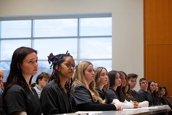 Twelve nursing students wearing dark gray scrubs sit together at a table in the College of Health Professions listening to their pledge ceremony.