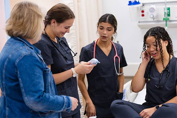 CHP Nursing Students 600x400 Dr. Suzanne Hakeos wearing a denim jacket stands with three nursing students wearing dark grey scrubs and stethoscopes. One student holds a thermometer that everyone is looking at.