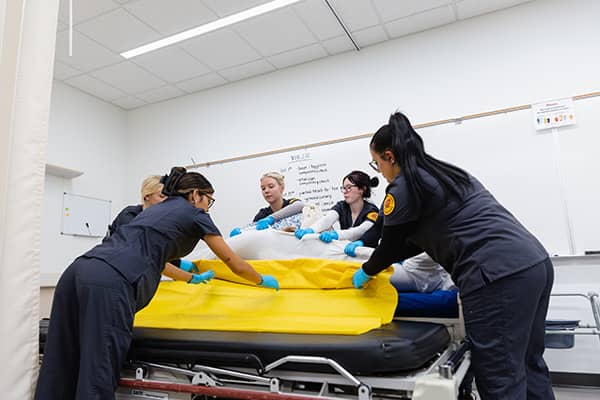 Five nursing students wearing dark grey CMU scrubs surround a hospital bed. Three students hold a white bedsheet upright from behind the bed while the other two students slide a yellow blanket underneath from the front of the bed.