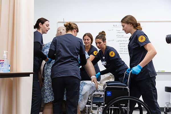 Nursing Transfer Simulation 600x400 Five nursing students wearing dark grey CMU scrubs surround a large patient mannequin. One student supports a wheelchair, two students hold the mannequin from the front while the last two support the mannequin from behind.