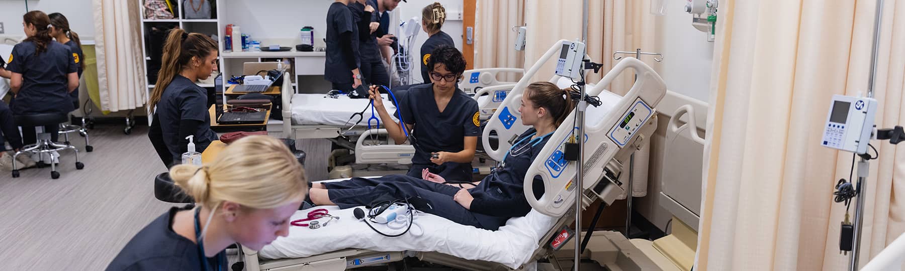 Nursing Students Vitals Simulation 1800x540 Ten nursing students dressed in dark gray scrubs practice taking vital signs in the simulation lab. One student sits at the front of the lab working on a laptop while others are standing or sitting in the background.