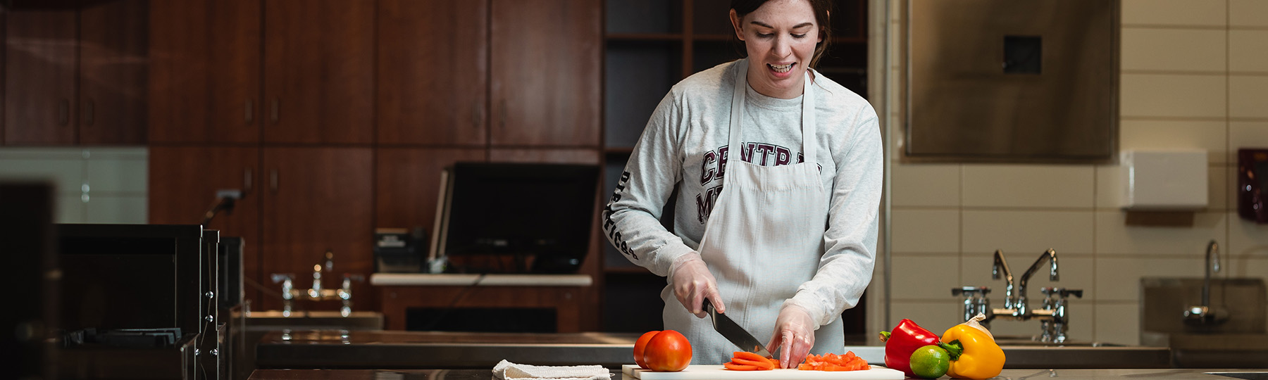 Student in a CMU shirt and apron slices vegetables on a cutting board in a campus kitchen.