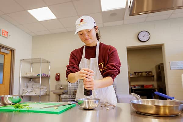 A student wearing a white CMU cap, white apron, and maroon sweatshirt stands behind a stainless steel table holding a pepper grinder above a stainless steel mixing bowl. A green cutting board, knife, and other stainless steel pans are also on the table.