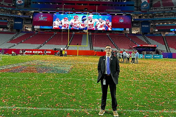 Sport Management student at Super Bowl LVII 600x400 Sport management student, Quintin Hunsinger, stands on the State Farm Stadium football field covered in red and yellow confetti with the Super Bowl 57 Champions, Kansas City Chiefs football players pictured on the jumbotron behind him in the endzone.