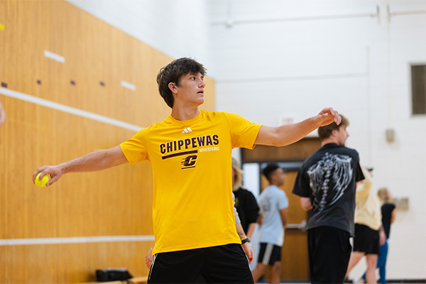 A physical education student wearing a gold Chippewas wrestling t-shirt stands in a T position preparing to throw a tennis ball held in his right hand.