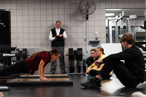 Physical Education Students in the Weight Room 600x400 A student in a maroon t-shirt demonstrates a push-up position while three other students sit on the floor and watch in a weight room. Dr. Rick Ferkel stands in the distance evaluating the demonstration.