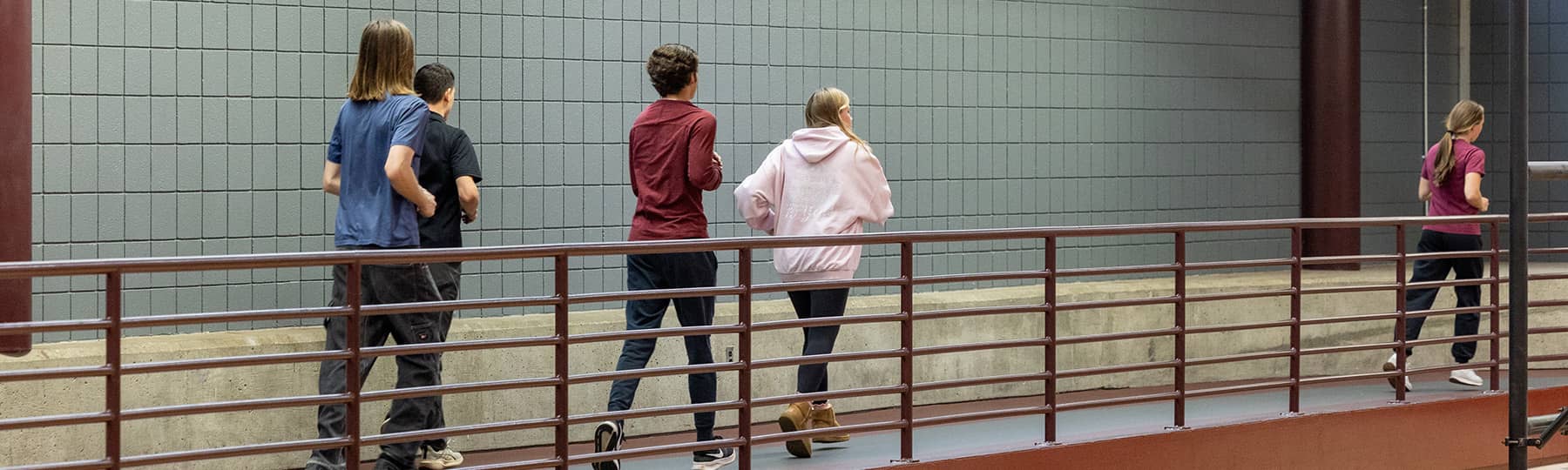 Health and Physical Education Class 1800x540 Five students jog behind a maroon metal fence on the indoor track in the Student Activity Center. One student is lengths ahead of the other four students.