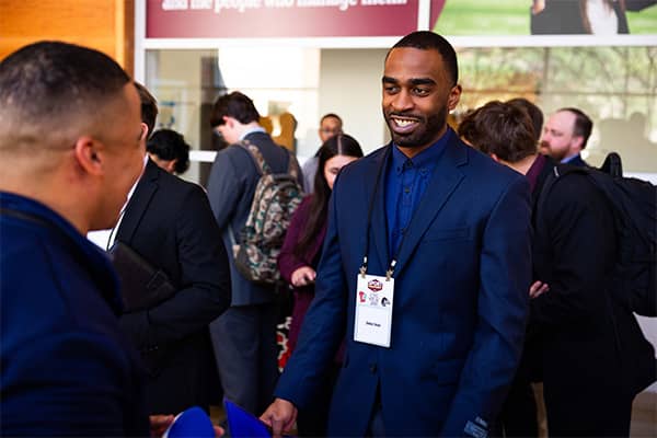 A man dressed in a blue suit stands smiling while looking at another man in a crowd of students at the sport management career fair in the College of Health Professions Building.
