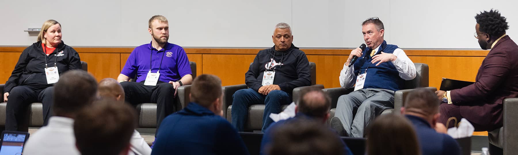 Sport Management Focus Group 1800x540 A woman and four men sit in chairs in front of an audience in an auditorium. One man is talking and holding a microphone while the other four are looking at him and listening.