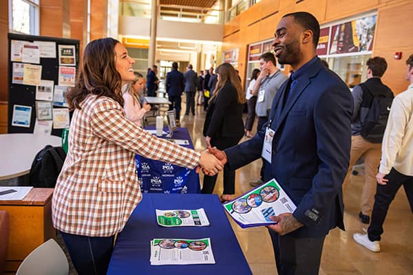 Sport Management Career Fair 600x400 A woman dressed in a brown and white checkered blazer reaches across a blue table to shake hands with a man dress in a navy blue suit. The man is holding papers in his left hand.