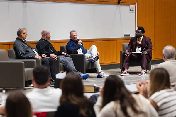 Four men sit in chairs in front of an audience in an auditorium. One man is talking and holding a microphone while the other three are looking at him and listening.