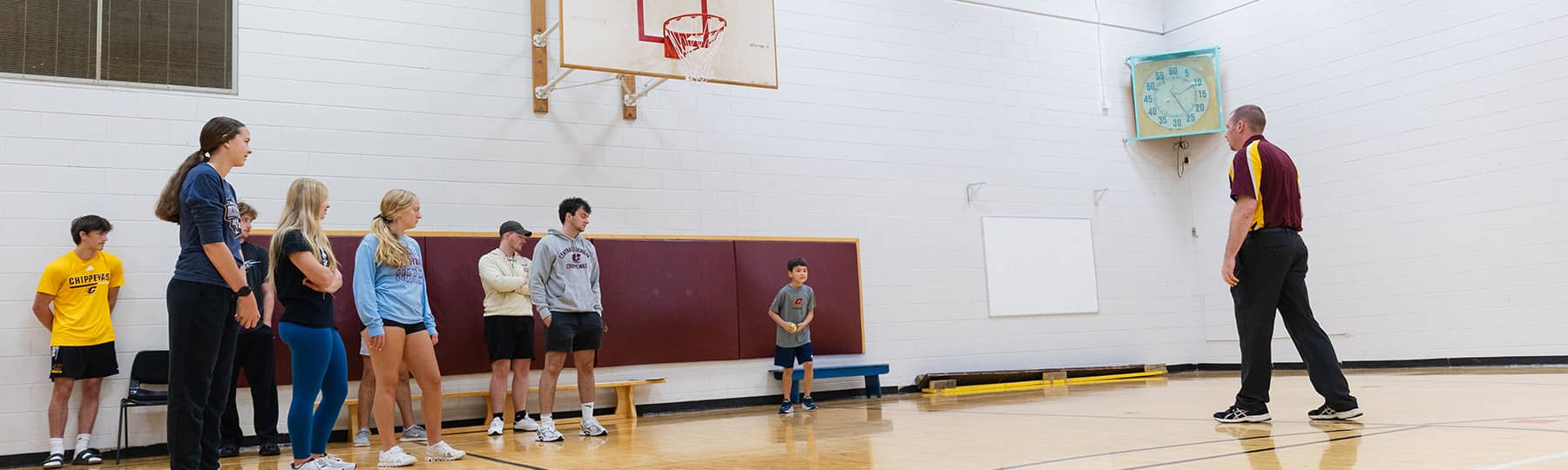 Dr. Kevin Fisher, wearing black pants and a maroon and gold polo, stands in front of eight students and a small young child in a gymnasium with a basketball hoop.