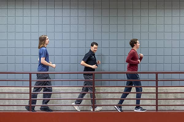 Three students walk briskly behind a maroon metal fence on the indoor track in the Student Activity Center.