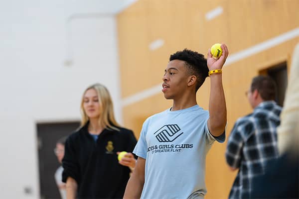 A physical education student wearing a light blue Boy and Girls Club of Greater Flint t-shirt stands and holds a tennis ball in his left hand with his left arm and elbow in a 90 degree position.
