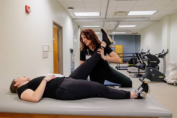 A physical therapy student dressed in black supports a patient's left leg in a raised position while the patient dressed in black lies flat on an examination table in the Carls Center Physical Therapy Clinic.