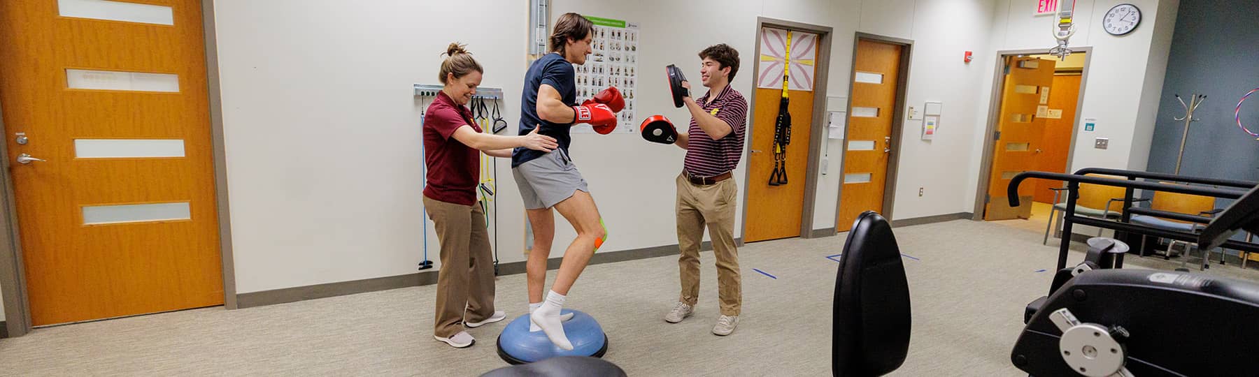 A patient wearing red boxing gloves balances on a BOSU ball while a Physical Therapist wearing a maroon polo and khaki pants holds his hips. A physical therapy student wearing a maroon polo and khaki pants stands in front of the patient  with boxing pads.