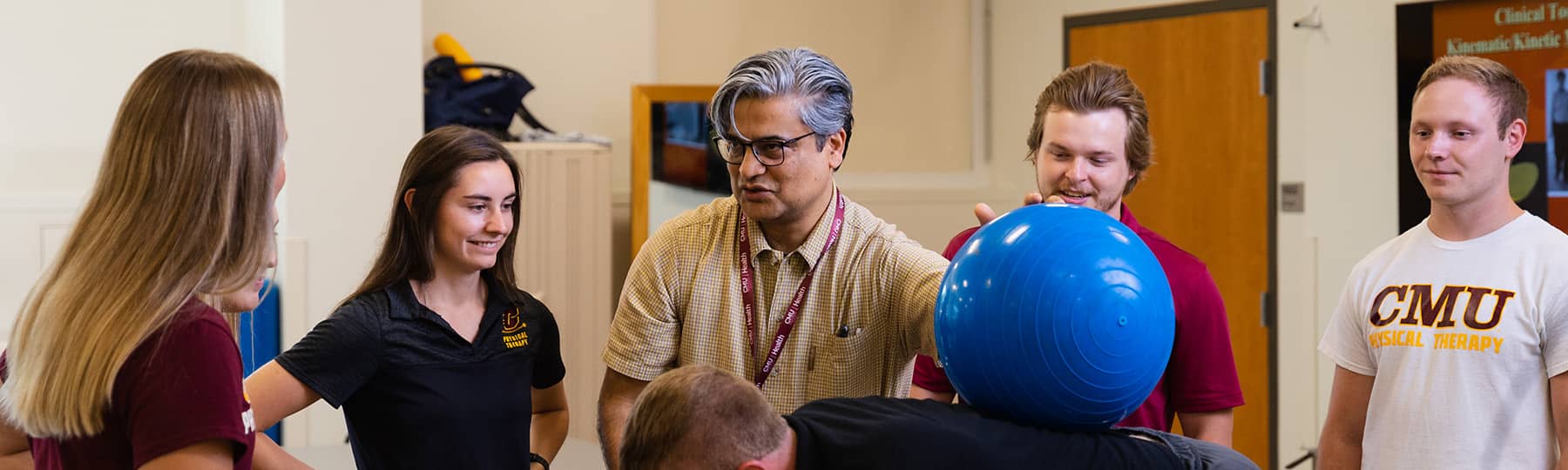Physical Therapy Lab Instruction 1800x540 Dr. Raju Chowdhary, wearing glasses and a tan polo, stands and talks to four Physical Therapy students while holding a bright blue exercise ball on an individual's back with his left hand.