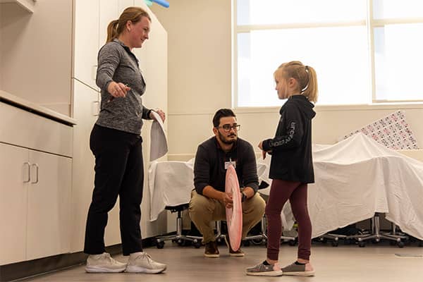 A Physical Therapy student dressed in black pants and a gray top stands in front of a young girl and gives her instruction for an exercise. A second Physical Therapy students squats down near the young girl while holding a pink disk in his hands.