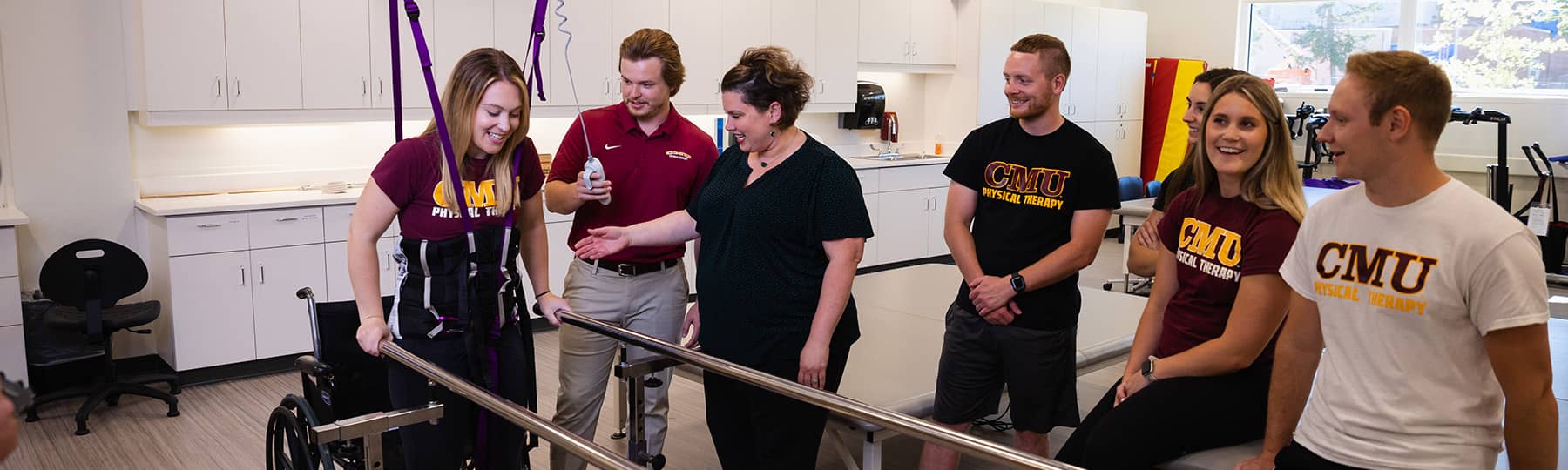 Dr. Rochelle Kopka, dressed in black, stands between four Physical Therapy students wearing CMU t-shirts. She instructs the students on training while having a student harnessed  in supports and standing between two parallel bars.
