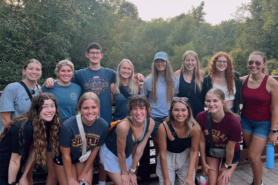A group of thirteen students from the Pre-Physical Therapy club at Central Michigan University pose for a group photo outside. They are all standing on a bridge smiling with trees and water behind them.