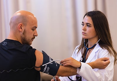 A physician assistant student in a white coat practices assessing blood pressure on a patient in a black t-shirt.