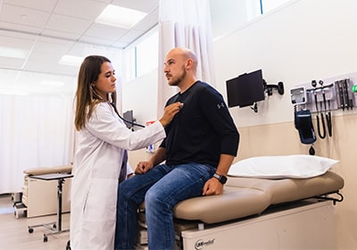 A physician assistant student in a white coat uses a stethoscope to listen to the heart of a patient in a black shirt.