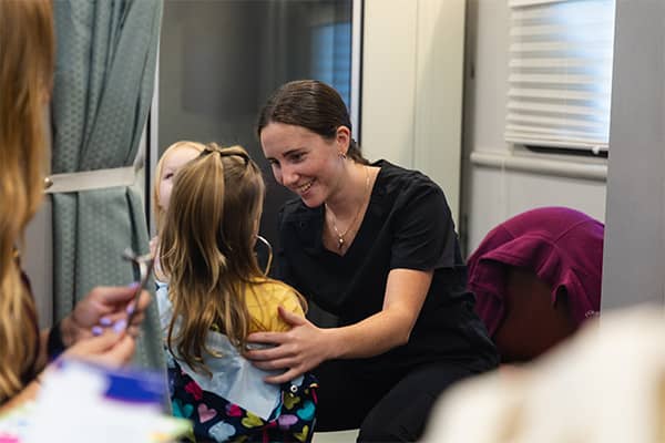A PA student in black scrubs smiles while sitting in front of a young child with her left hand on the child's upper back. The child has long brown hair and is using a stethoscope to listen to her heart in the Mobile Health Central bus.