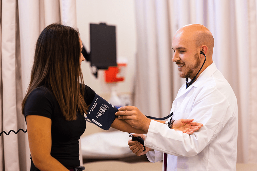 A male doctor is smiling and checking a female patient's blood pressure with her left arm facing towards him.