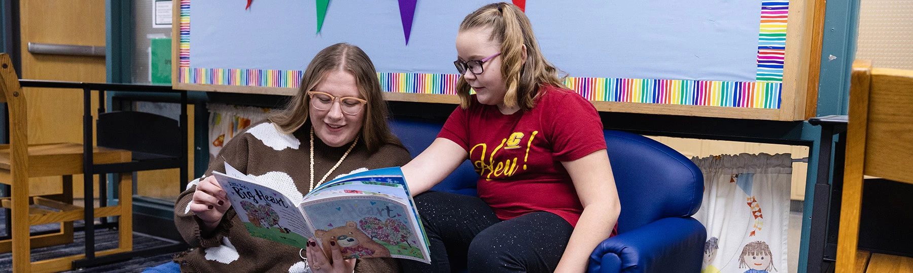 A female teacher with glasses holding a story book sits next to a female elementary student in glasses who is pointing to the words on the story book's page as she reads.