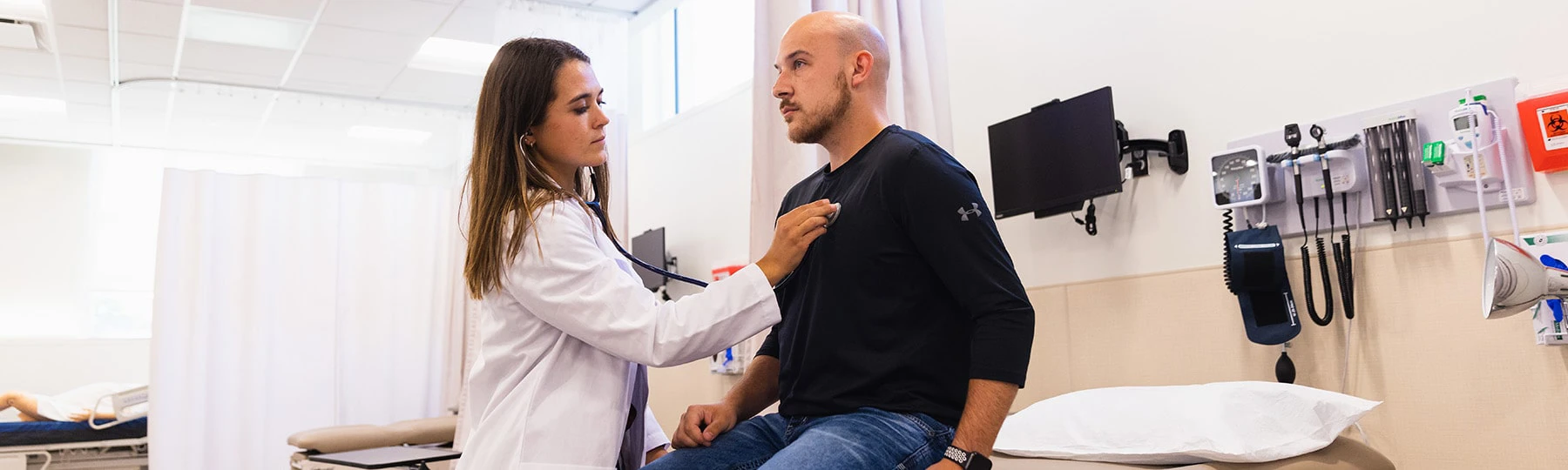 A physician assistant student in a white coat uses a stethoscope to listen to the heart of a patient in a black shirt.