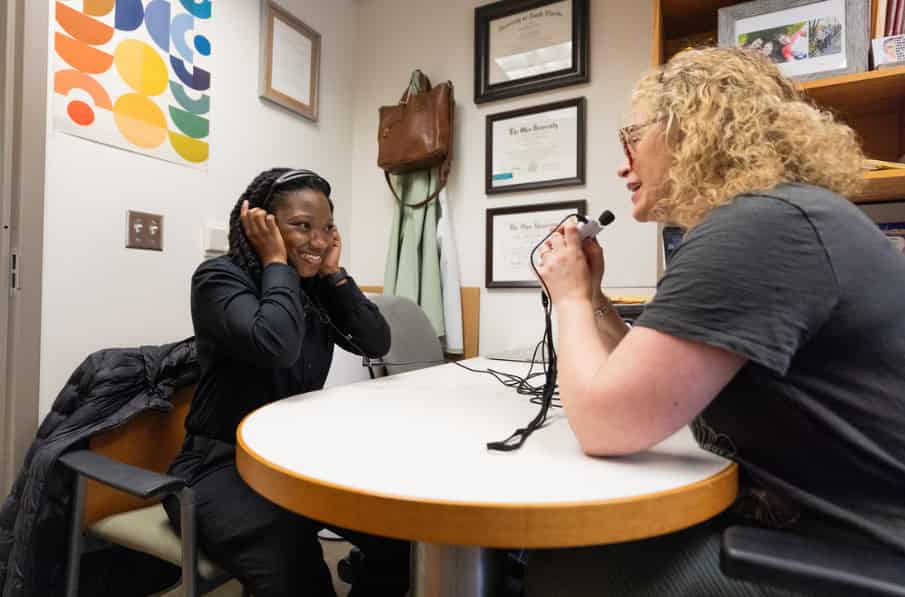 A speech language pathologist student wears headphones as a professor speaks into a microphone. The student is sitting at a desk across from the professor inside of an office.