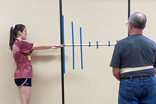 A physical therapy student stands in front of a measuring device taped to the wall with her arms extended while an older male watches her demonstration.