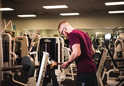 An exercise science student in a maroon t-shirt stands behind a bench press and spots on individual pressing up a weighted barbell.