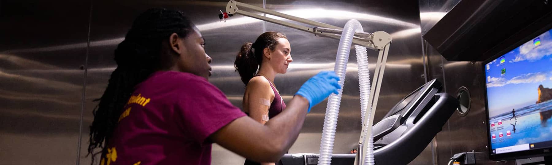 Exercise Physiology Research 1800x540 A student dressed in shorts and a tank top stands on a treadmill looking at a computer screen while a second student wearing a maroon t-shirt reaches for an opaque hose while standing near the treadmill and computer.