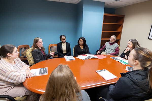 Seven females and one male sit at a round conference table discussing public health topics. There are papers spread around the table in front of individuals.