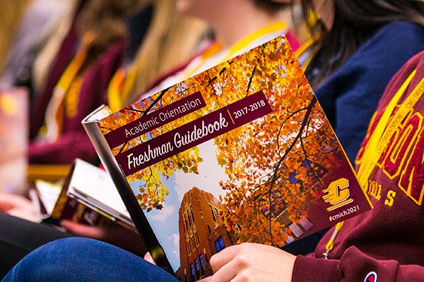 A student holds a booklet titled Academic Orientation Freshman Guide 2017-2018. The Cover features Warriner Hall framed by orange autumn leaves with maroon and gold design elements and Action C logo at bottom corner.