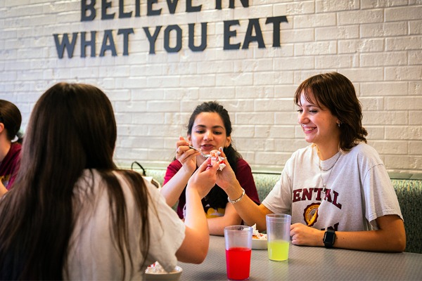 Three young women smiling and having a celebratory drink toast in a casual dining setting with a brick wall bearing the text BELIEVE IN WHAT YOU EAT